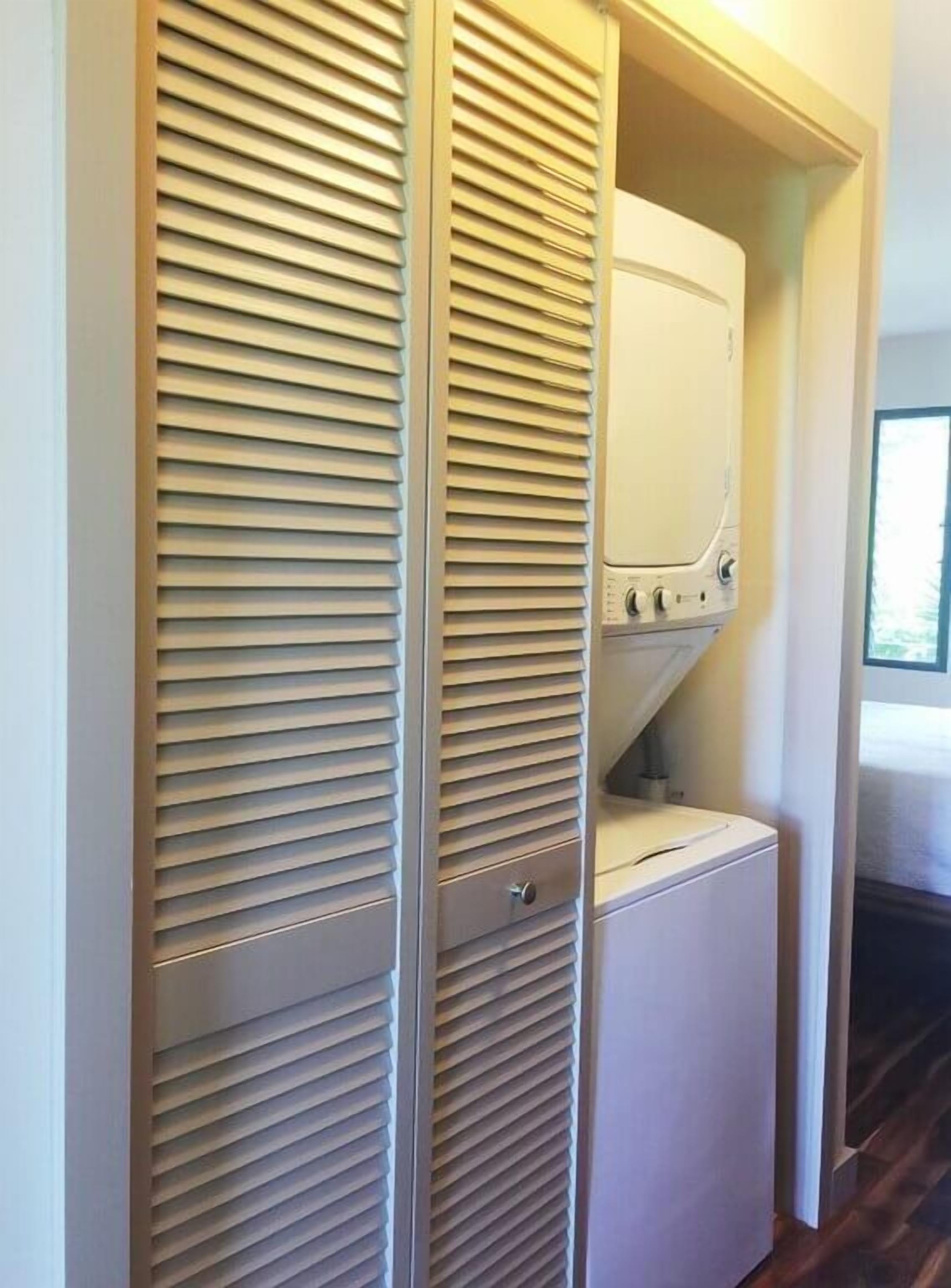 A laundry room with a front-loading washer and dryer, a wooden door with horizontal slats, and a window providing natural light.