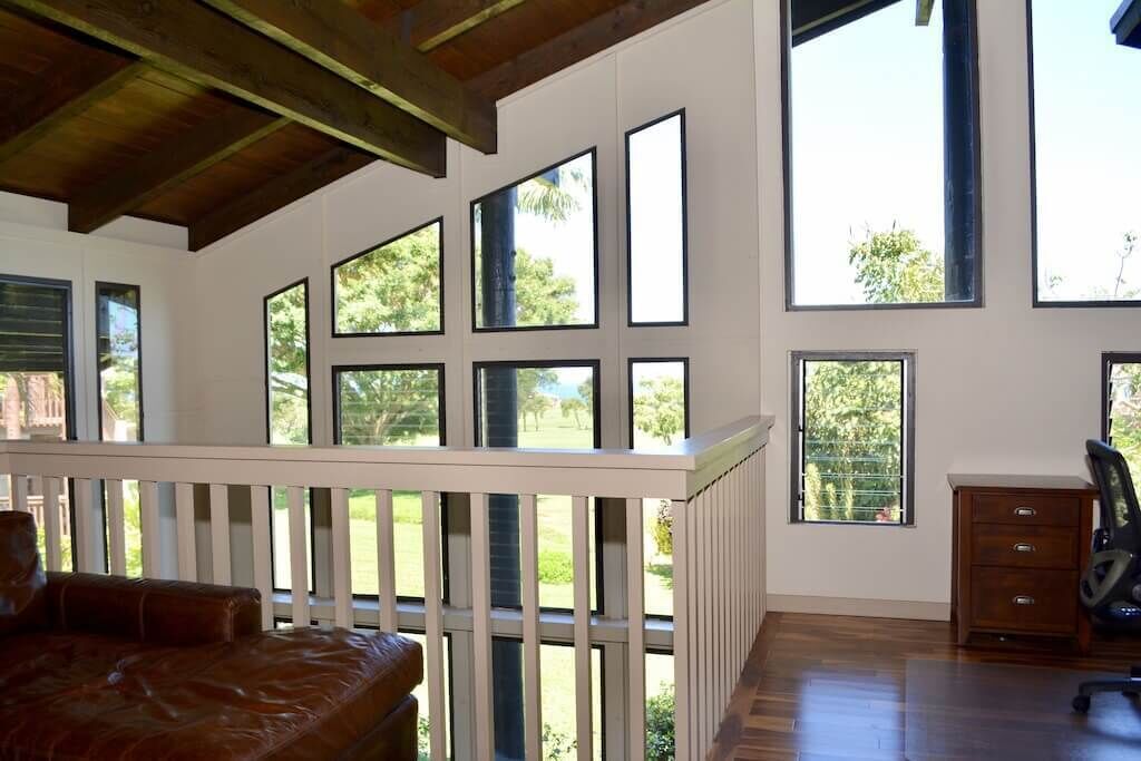 Une pièce dotée de grandes fenêtres, d’un plafond en bois, d’une rambarde blanche, d’un canapé en cuir marron et d’un bureau en bois avec des tiroirs.