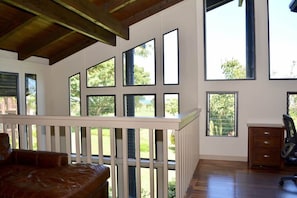 A room with large windows, a wooden ceiling, a white railing, a brown leather sofa, and a wooden desk with drawers. - Beautiful Tropical Getaway - Pool - Grill & More (Princeville)