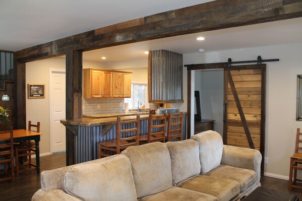 A cozy living room with a large beige sofa, wooden chairs, and a kitchen area with wooden cabinets and a bar.