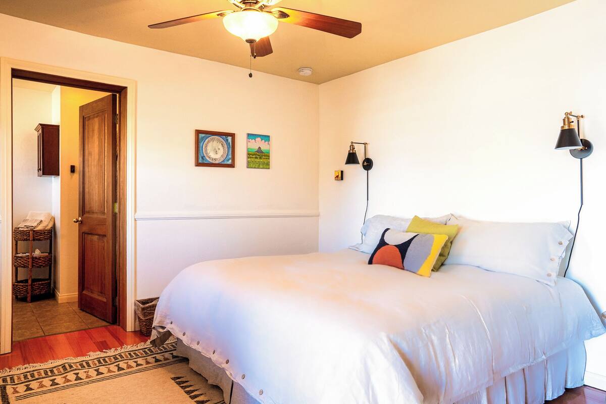 A bedroom with a bed, a ceiling fan, a wall-mounted lamp, and a door leading to another room.