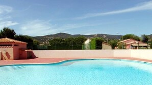 A swimming pool with a clear blue water surface, surrounded by a low wall and a paved deck. In the background, there are buildings with red roofs and a mountainous landscape. - Cozy House with Pool & Tennis, Near Beach, Sleeps 6 - Bormes-les-Mimosas (Bormes-les-Mimosas)