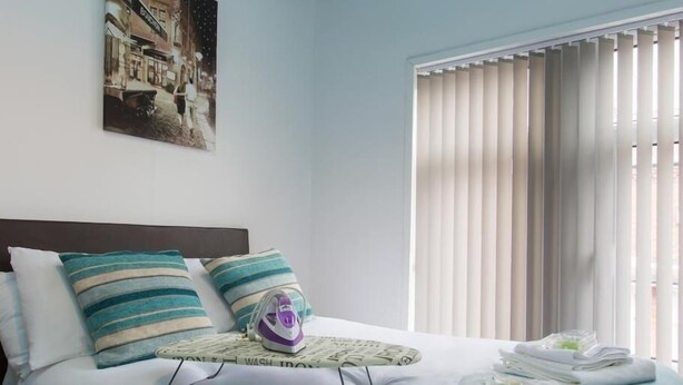 A hotel room with a bed, pillows, a small ironing board, and a window with blinds. - Townhouse @ Westminster St (Crewe)