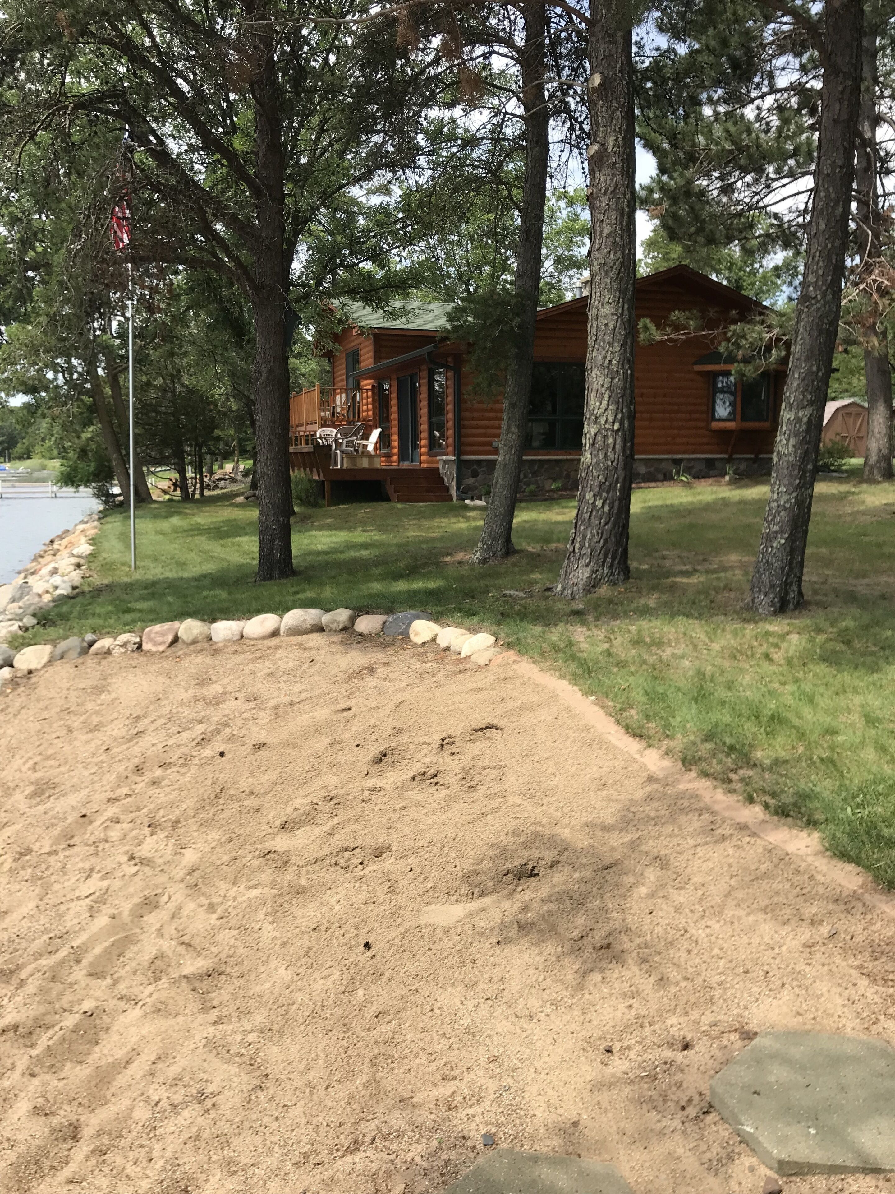 A cabin with a deck and outdoor seating, surrounded by trees and a sandy area.