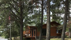A cabin with a deck and outdoor seating, surrounded by trees and a sandy area.