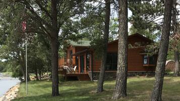 A cabin with a deck and outdoor seating, surrounded by trees and a sandy area.