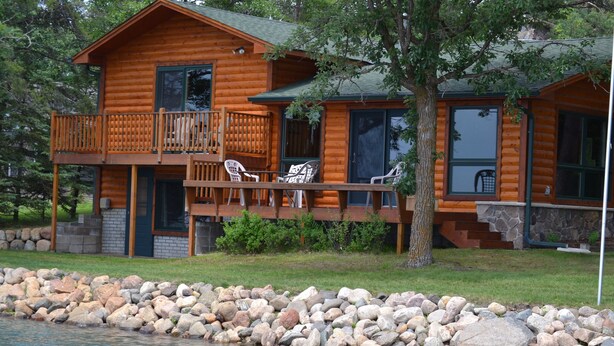 A log cabin with a deck, outdoor chairs, and a stone-lined water feature.