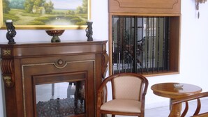 A room with a wooden fireplace, a chair, a table, and a window with bars. - Al Sakhra Hotel (Bhamdoun)