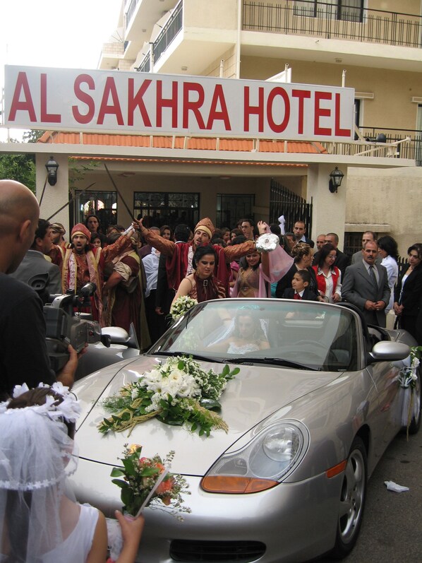 Uma procissão de casamento em frente ao Hotel Al Sakhra, com convidados e um carro decorado.