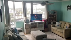 Ein Wohnzimmer mit Fernseher, Sofa, Hocker und Esstisch.