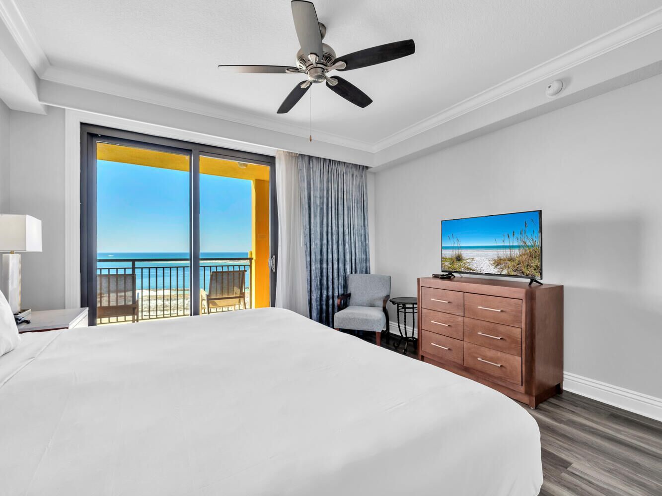 Habitación de hotel con una cama grande, ventilador de techo, televisor sobre una cómoda de madera, una silla y un balcón con vista a la playa.