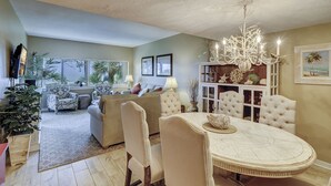 A dining area with a round table and chairs, a chandelier, and a view of the living room with a sofa and a television. - 464 Captains Walk - 2 bedroom, 2 bath villa convenient to the beach! (Hilton Head Island)