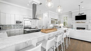Una cocina moderna con armarios blancos, una isla central con taburetes de bar, horno empotrado y un televisor grande montado sobre la chimenea.