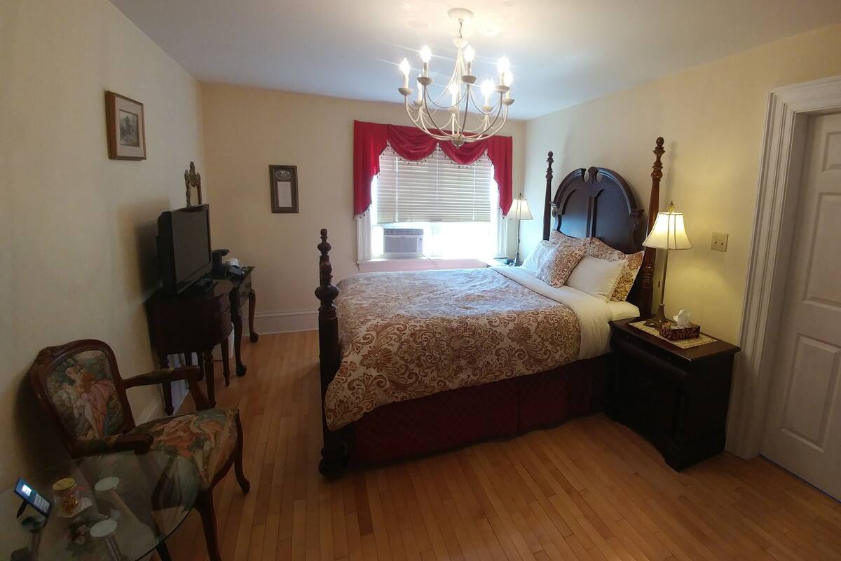A bedroom with a four-poster bed, a chandelier, a bedside table with a lamp, a chair, and a door.