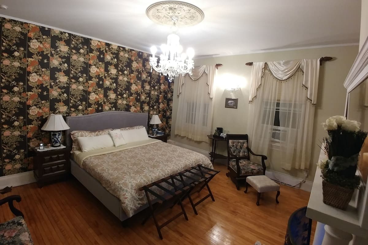 A bedroom with a floral wallpaper, a chandelier, a bed with a patterned bedspread, a wooden bench, and a chair with a floral cushion.