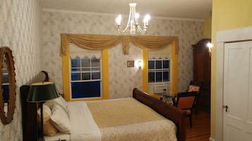 A bedroom with a large bed, two windows with curtains, a chandelier, a lamp, and a mirror.