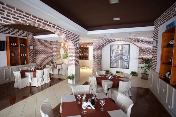 A restaurant with brick walls, white tables, and chairs. There are shelves with bottles and a painting on the wall.