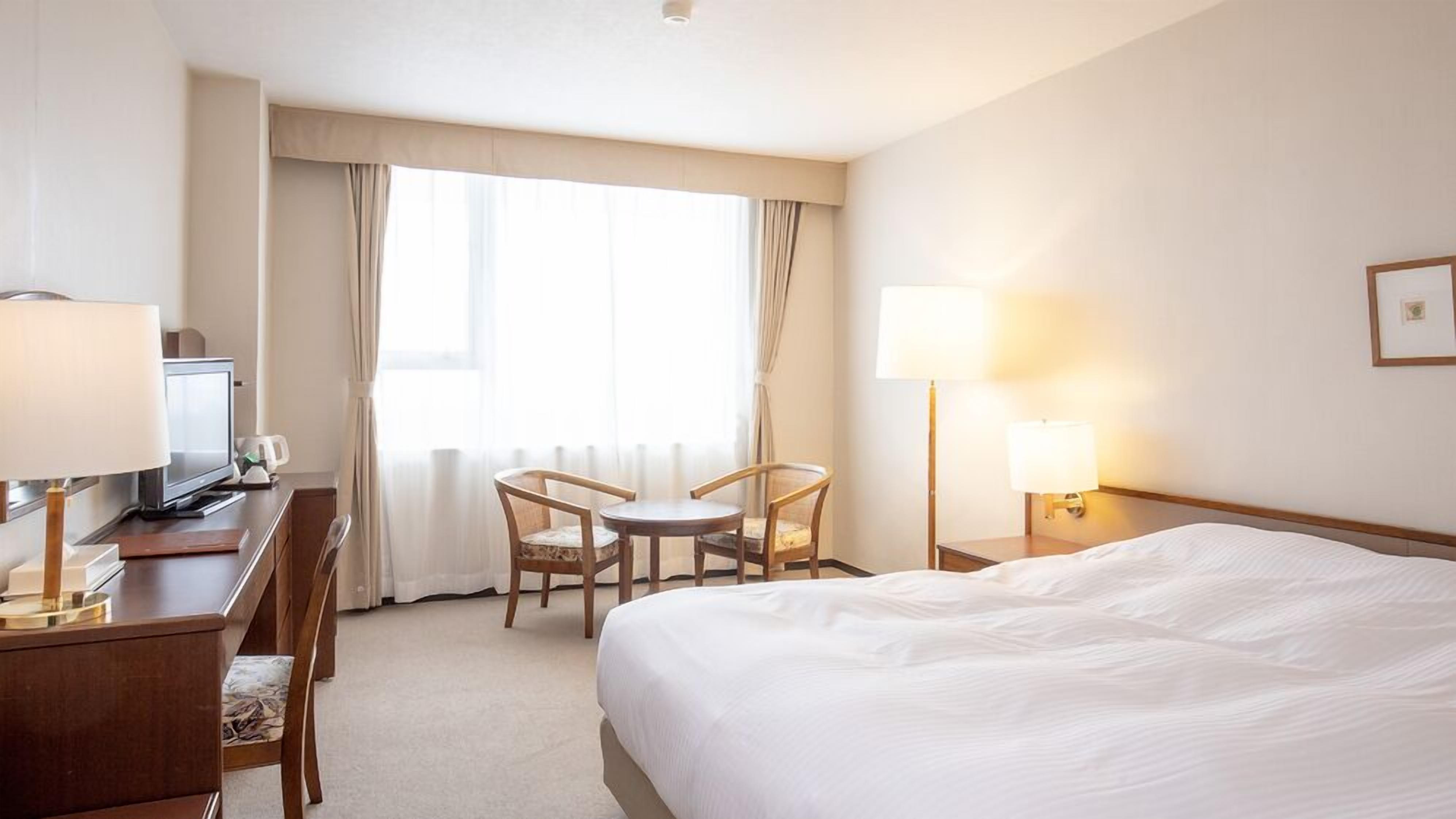 A hotel room with a bed, desk, chair, TV, and lamps.
