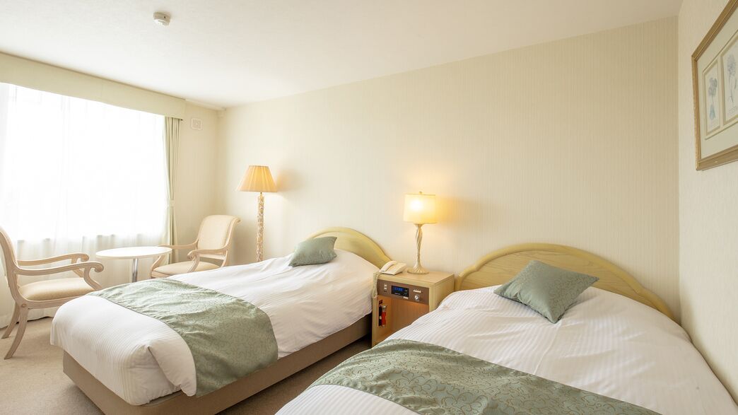 A hotel room with two beds, a chair, a small table, and lamps.