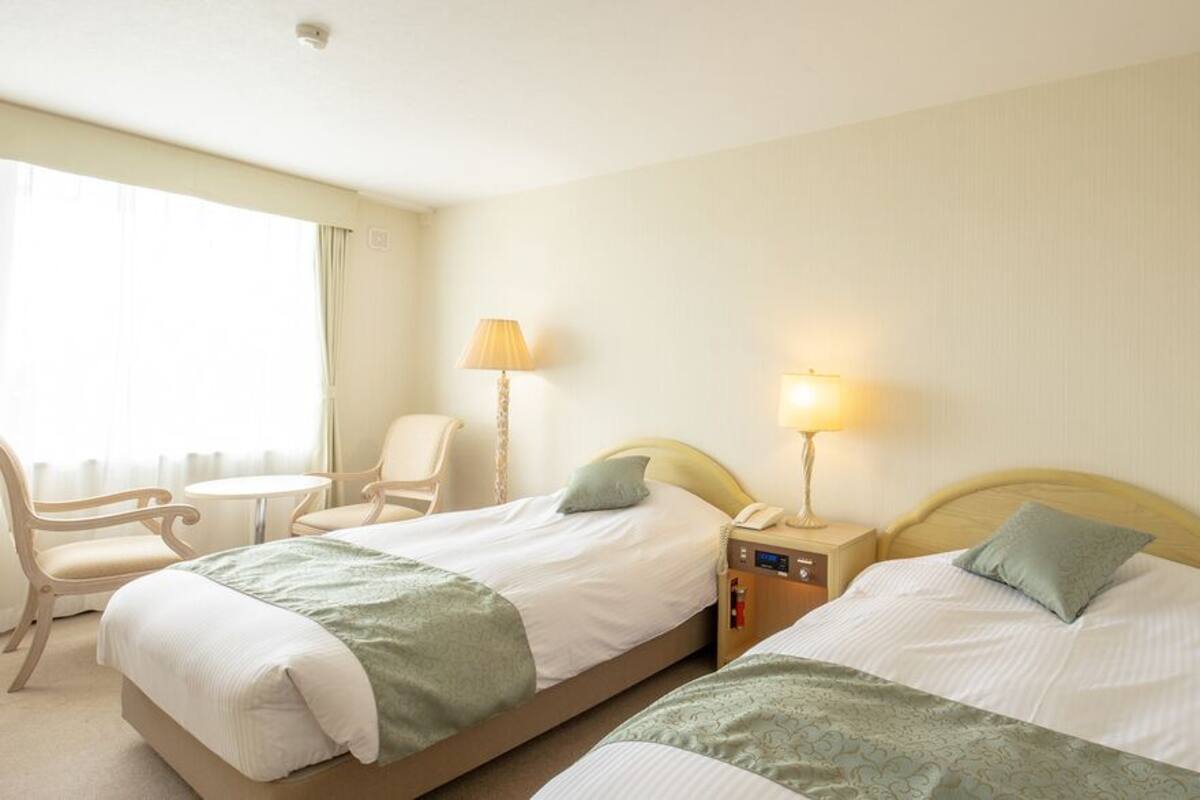 A hotel room with two beds, a chair, a small table, and lamps.