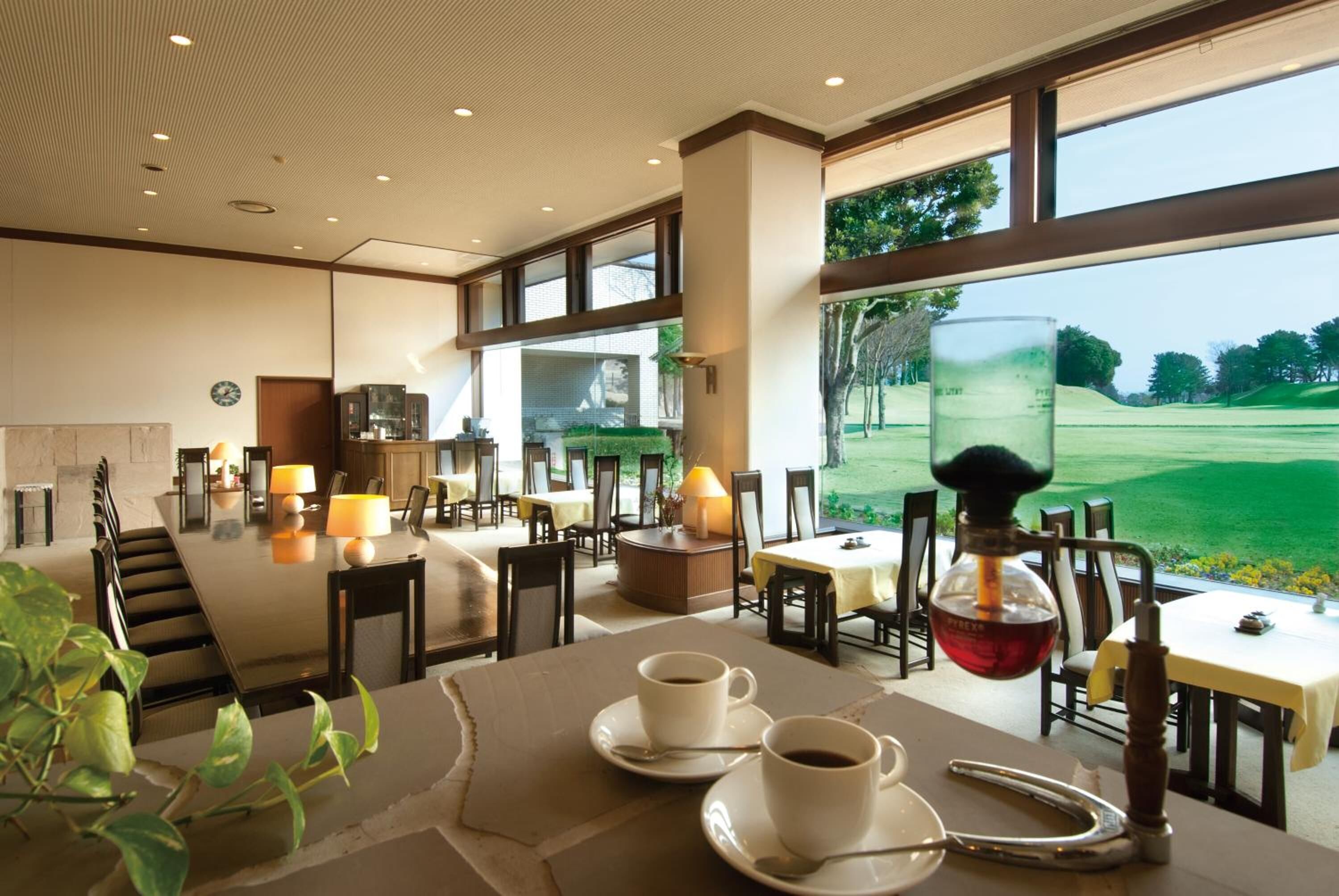 A spacious indoor dining area with large windows offering a view of a golf course. Tables are set with white plates and cups, and there are several lamps providing lighting.