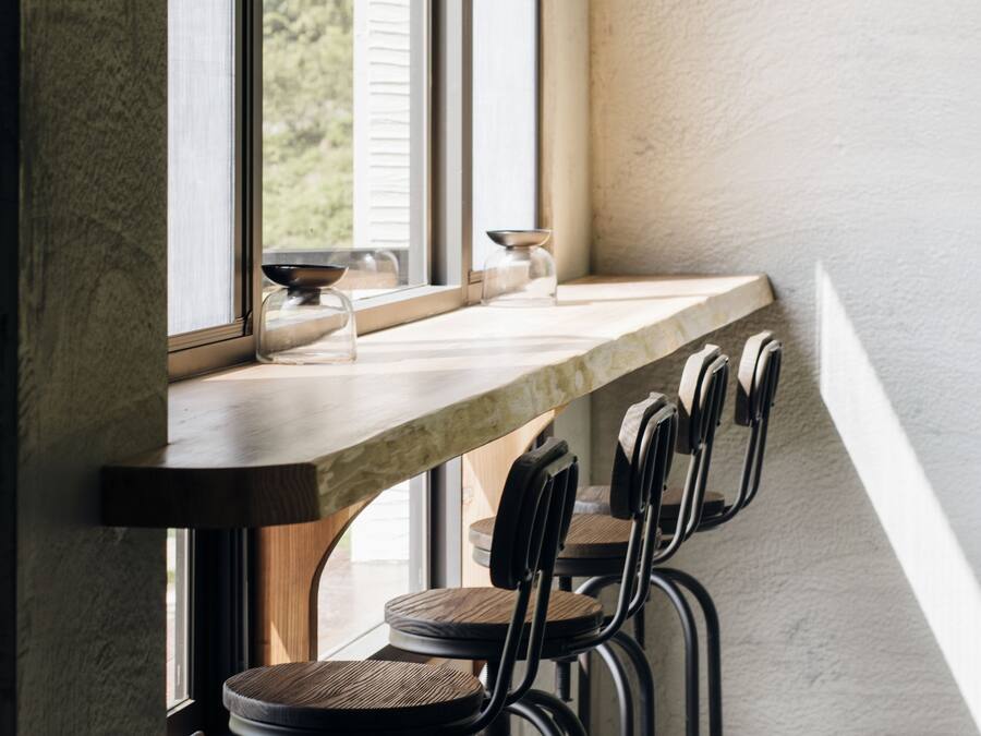 A modern kitchen with a long countertop, black bar stools, and a view of greenery outside.