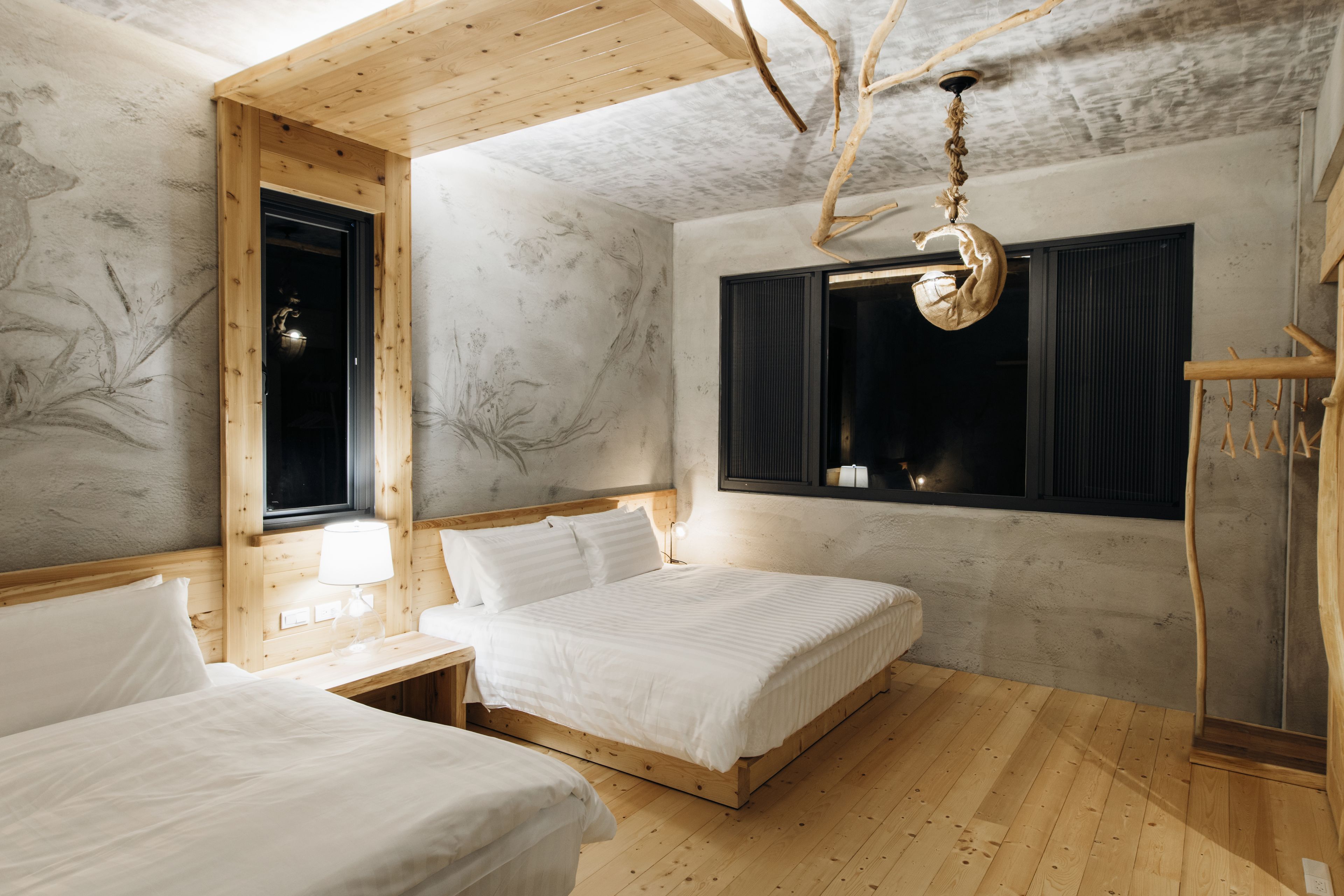 A room with two beds, wooden flooring, a window with curtains, a hanging light fixture, and a wall with a tree mural.