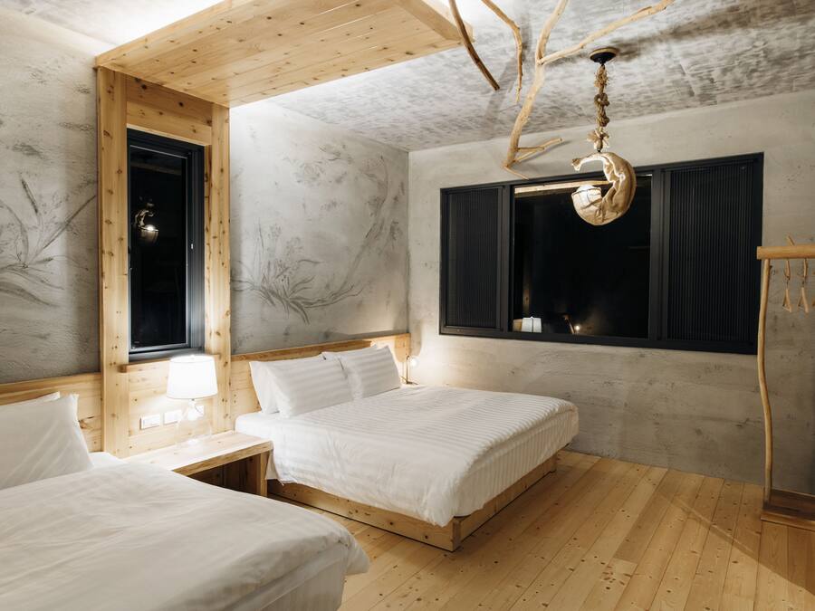 A room with two beds, wooden flooring, a window with curtains, a hanging light fixture, and a wall with a tree mural.