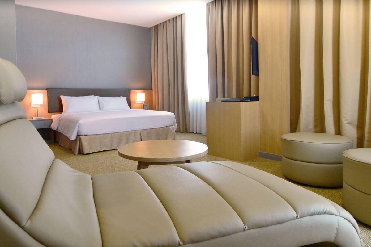 Ein Hotelzimmer mit einem großen Bett, zwei Nachttischen mit Lampen, einem Sofa, einem kleinen runden Tisch und einem an der Wand befestigten Fernseher.