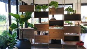 A modern interior with a wooden shelving unit holding books and plants, a large potted plant, and a view of the outdoors. - Hotel Everyday Life (Hengchun)