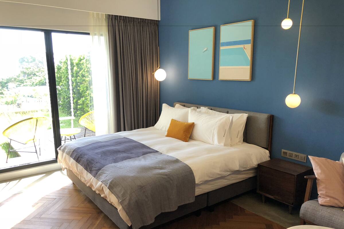 A modern bedroom with a large bed, a nightstand, a chair, and a view of the outdoors.