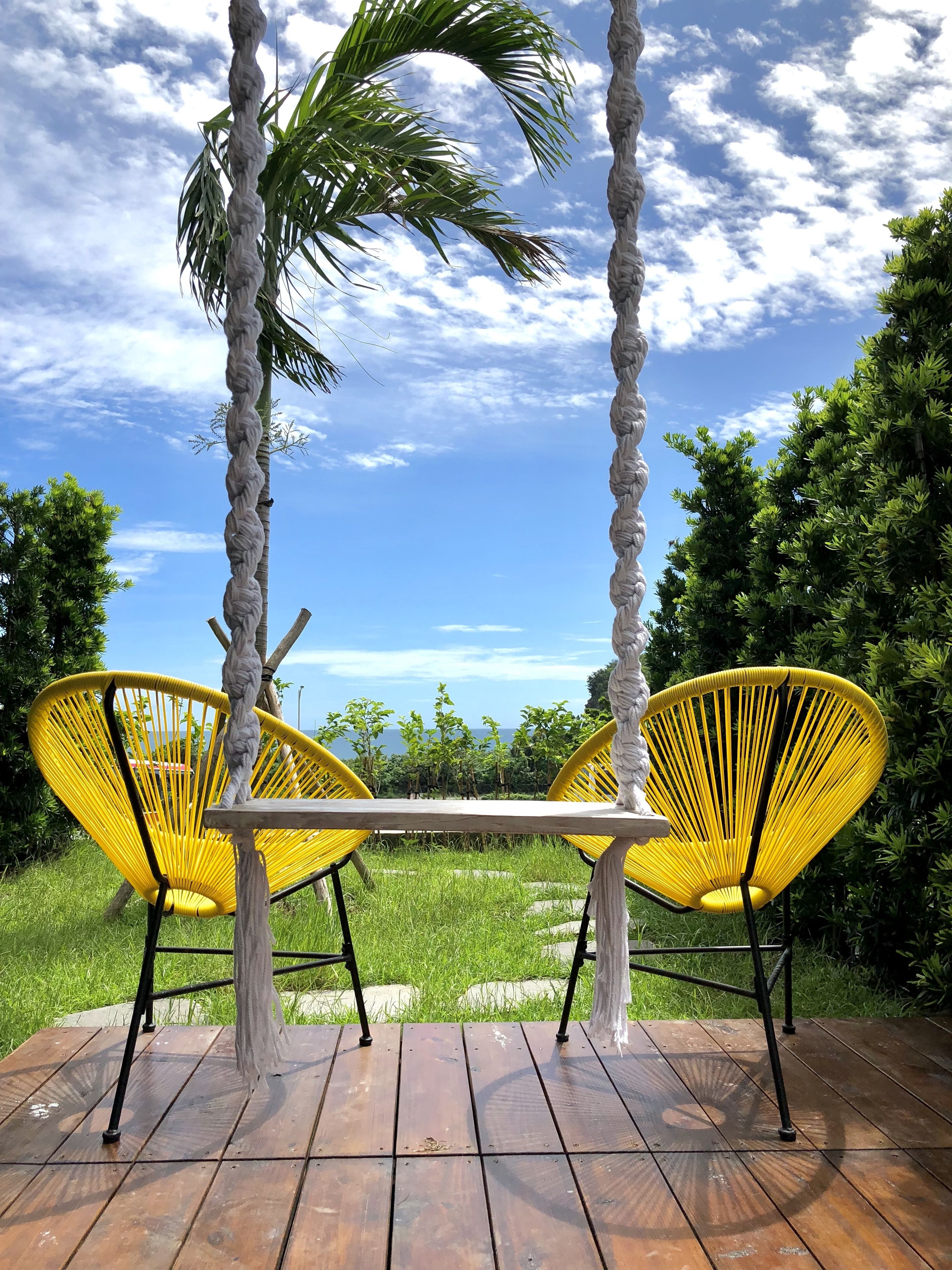 A wooden deck with two yellow chairs and a table, surrounded by palm trees and greenery, overlooking a landscape.