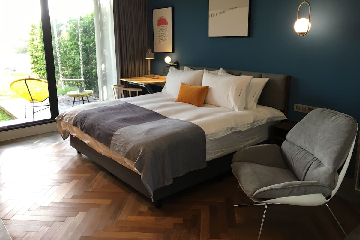 A modern bedroom with a large bed, a desk, and a chair. There is a window with a view of greenery outside.