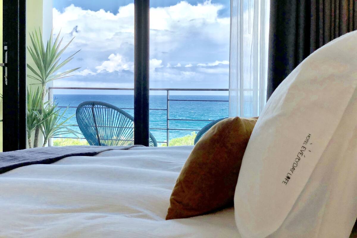 A hotel room with a bed, a view of the ocean, a chair, and a window with curtains.