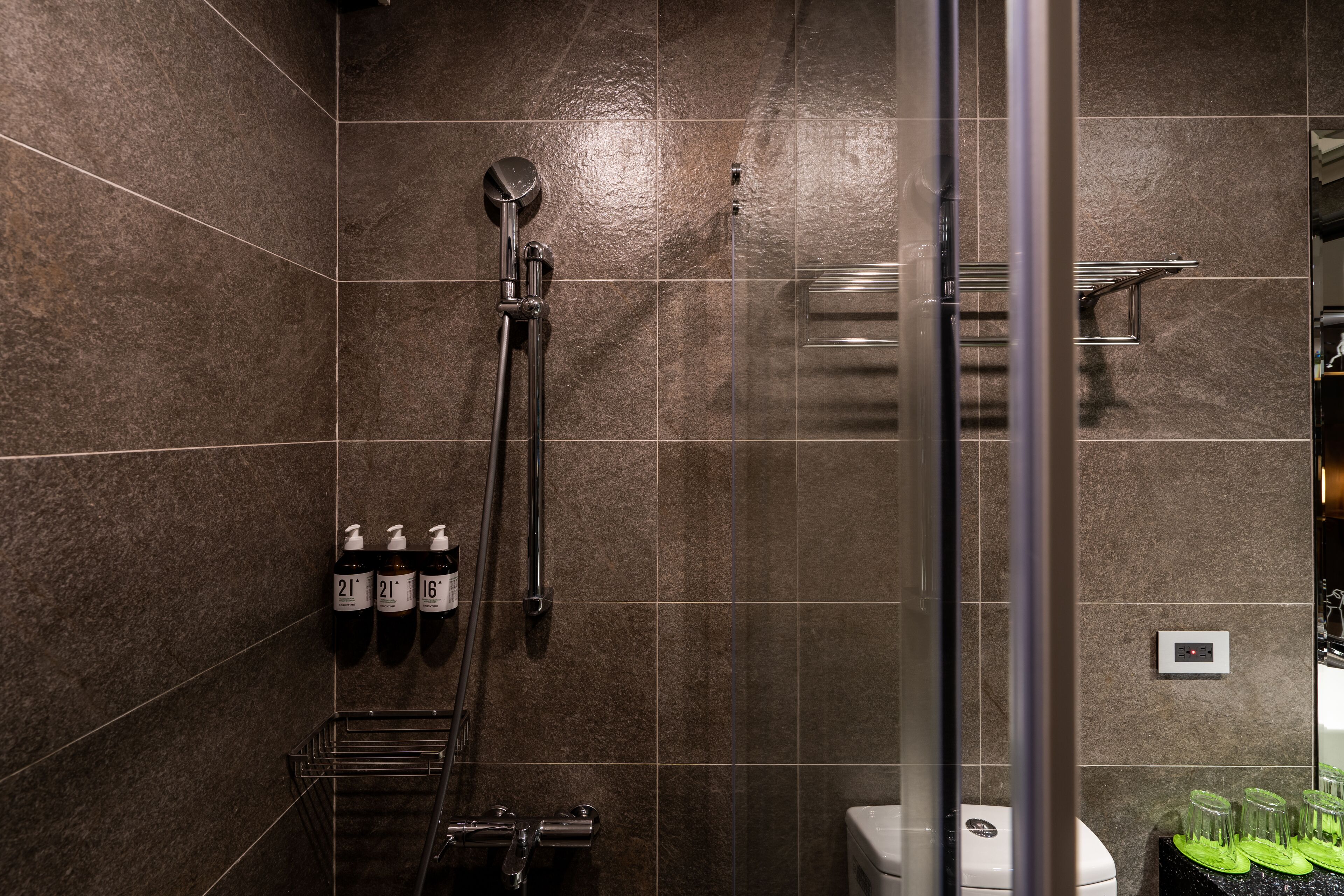 A modern shower area with a glass door, a handheld showerhead, and a wall-mounted faucet. There are three bottles of shampoo and conditioner on the wall.