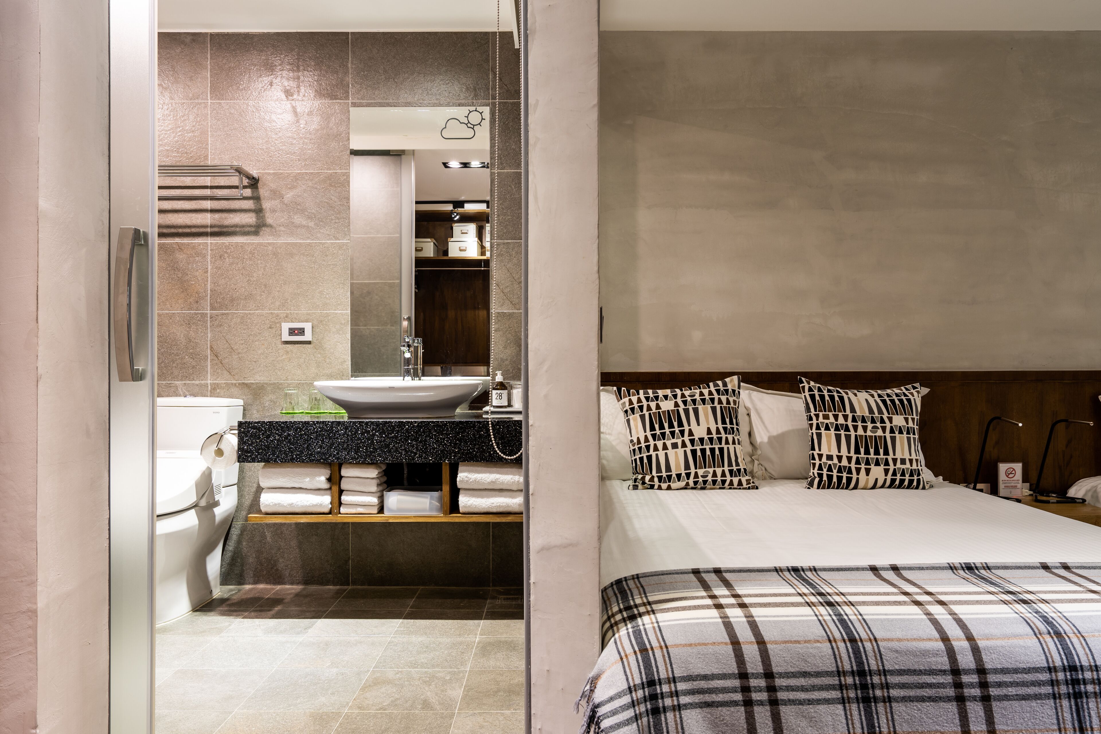 A modern bathroom with a sink, toilet, and towel rack. The room features a bed with plaid bedding and patterned pillows.