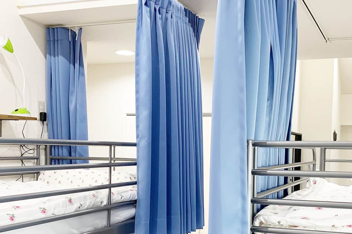 A dormitory room with bunk beds, blue curtains, a desk with a lamp, and a wall socket.