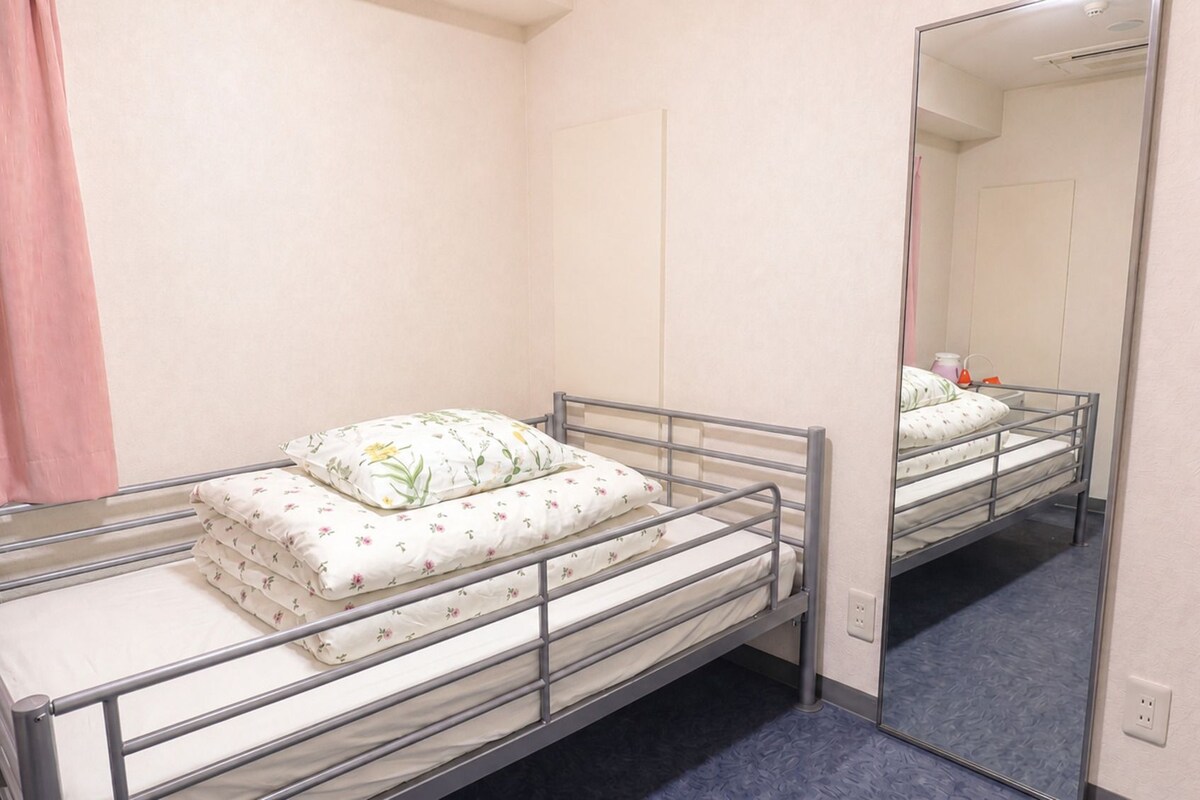 A single metal-framed bed with floral bedding, a pink curtain, and a mirror reflecting the room.