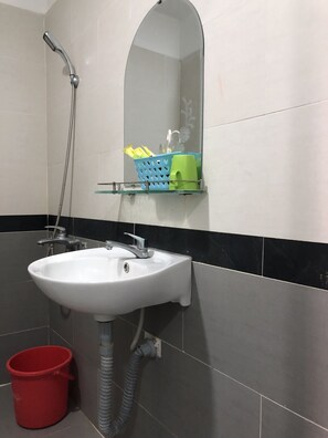 A bathroom with a white sink, a glass shelf with toiletries, a red bucket, and a shower. - Gold Hotel (Ho Chi Minh City)