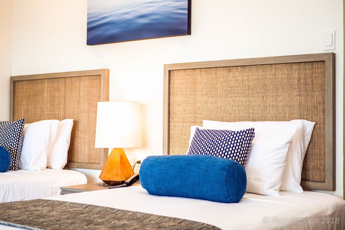A hotel room with two beds, each with a headboard, white bedding, and blue and patterned pillows. A lamp is on the nightstand between the beds.