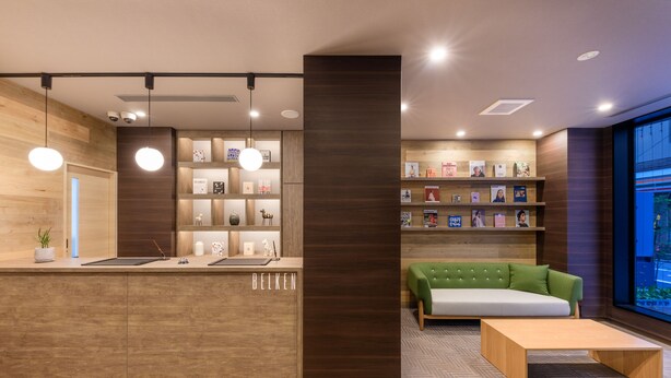 A reception area with a wooden reception desk, a green sofa, and a bookshelf with books and decorative items. - Belken Hotel Kanda (Tokyo)