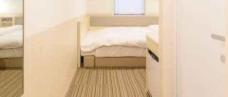 A narrow hotel room with a single bed, a small window with curtains, a mirror, and a carpeted floor.