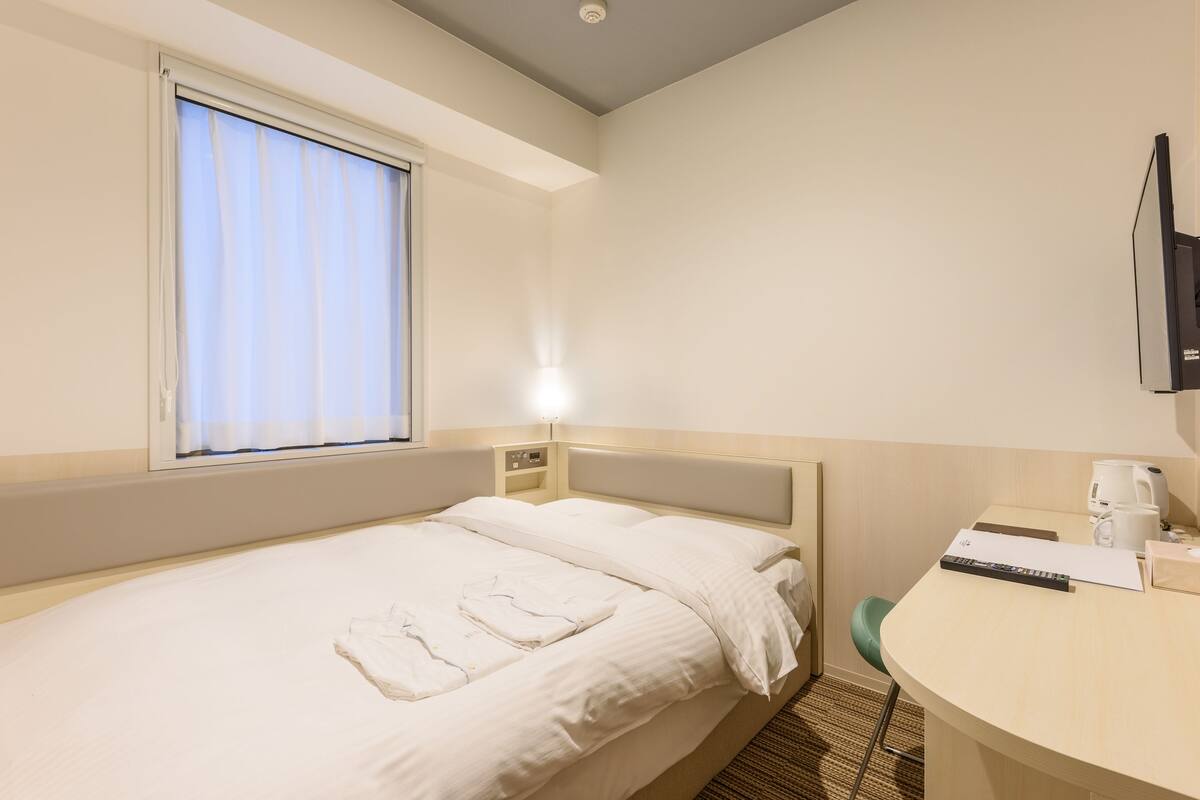 A hotel room with a single bed, a bedside table with a lamp, a desk with a chair, and a window with curtains.