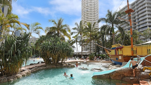 Ein Poolbereich mit Wasserrutsche, Palmen und einem großen Gebäude im Hintergrund.