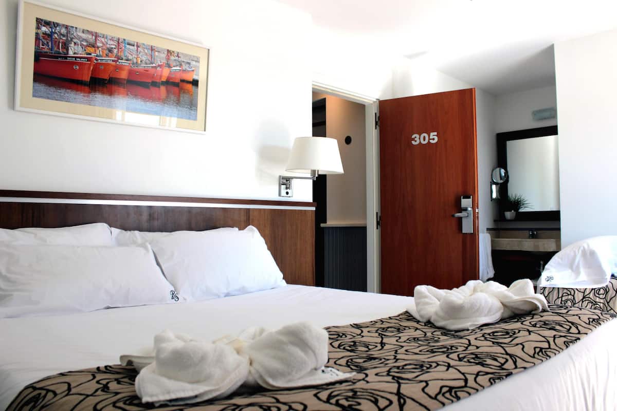 A hotel room with a large bed, a wooden door labeled 305, a painting of boats on the wall, and a bedside lamp.