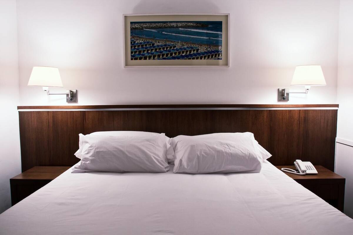 A hotel room with a wooden headboard, two bedside lamps, a phone on the nightstand, and a framed picture of a beach scene on the wall.
