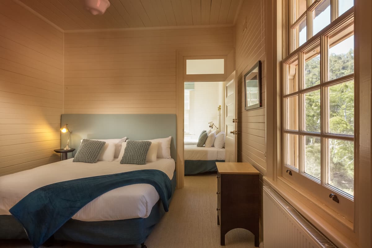 A bedroom with two beds, a bedside table, a lamp, and a window with a view of greenery.