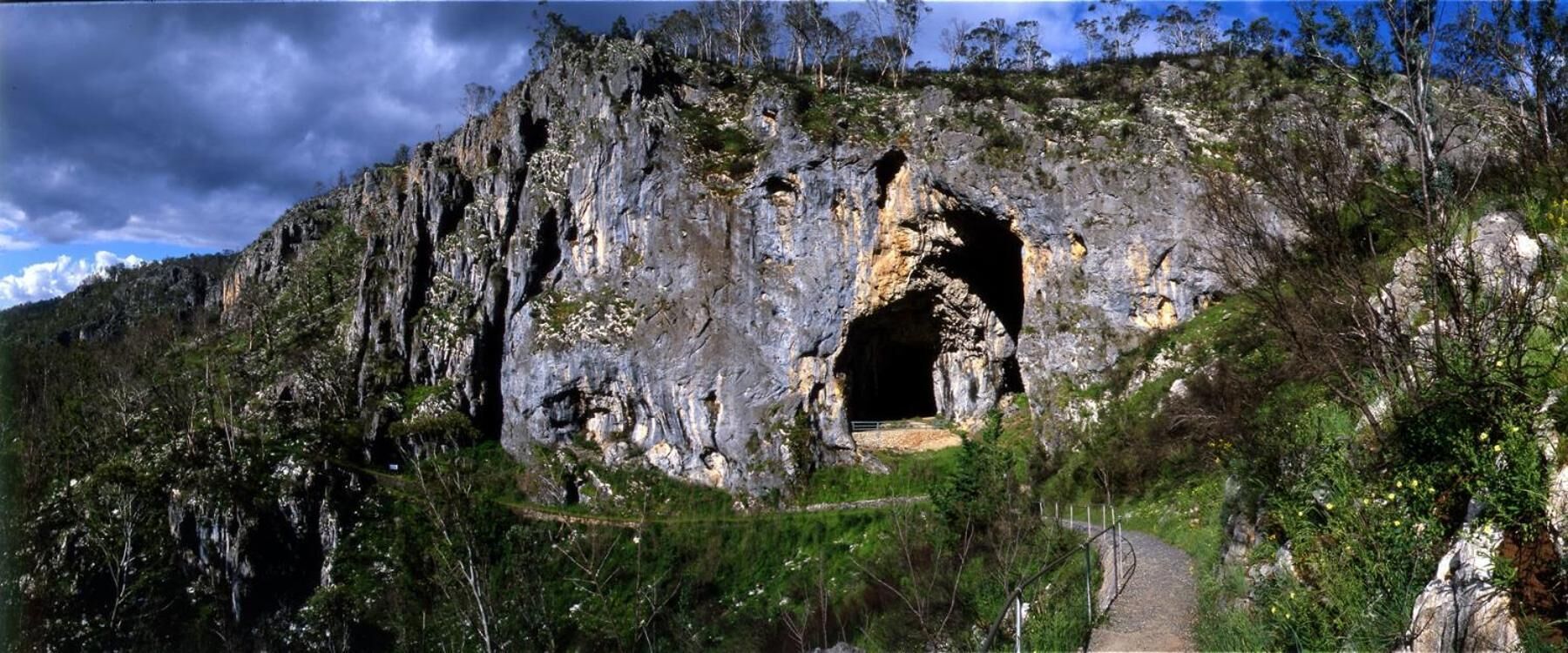 A natural rock formation with a cave entrance, surrounded by greenery and a pathway leading to it.