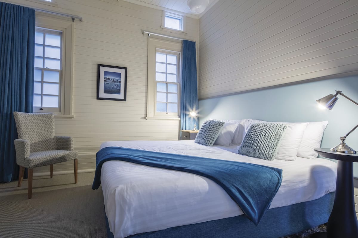 A neatly arranged bedroom with a large bed, a chair, a bedside table, and a framed picture on the wall.