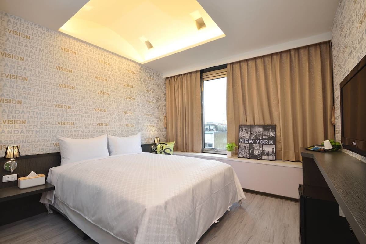 Habitación de hotel con una cama grande, una mesita de noche con lámpara, una ventana con cortinas y una pared con texto repetido.